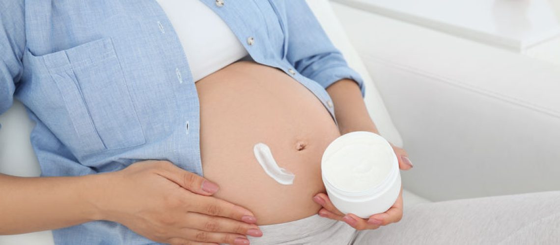Pregnant woman applying body cream on belly at home, closeup